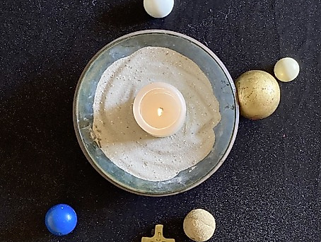 Kerze in einer Sandschale rundherum ist angeordnet ein Kreuz und verschiedenfarbige Perlen