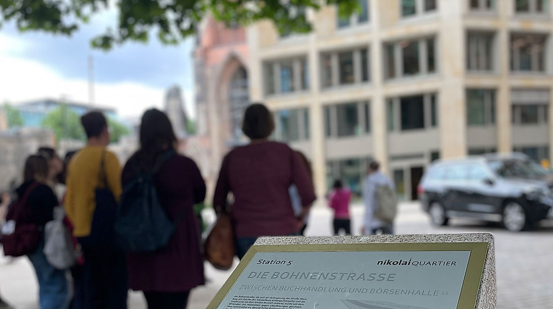 Infotafel Bohnenstrasse mit tastbarer Karte in Hamburg, dahinter eine Gruppe von Menschen und modernes Gebäude 