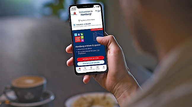 Eine Person sitzt in einem Café mit Cappuccino und Keksen auf dem Tisch und hält ein Smartphone in der Hand. Auf dem Display ist die mobile Hamburg CARD mit Rabattinformationen und Ticketsichtbarkeit zu sehen. Die Szene symbolisiert modernes, unkompliziertes Reisen mit allen Vorteilen digital zur Hand – jederzeit einsatzbereit.