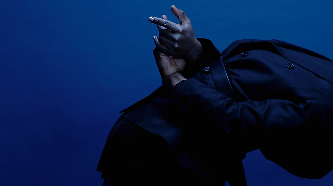 Ein*e Performer*in ist von der Seite zu sehen und steht nach vorne gebeugt, der schwarze Kaputzenpulli fällt über den Kopf. Die Hände sind hinter dem Rücken und ausgestreckt.