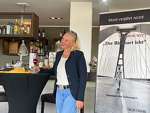 Die Autorin Nadine Witt steht neben einem Roll-up mit dem Cover ihres Buches „Der Blaubart lebt“ – Stimmen der Zeitzeugen. Sie blickt leicht zur Seite und trägt einen Ausdruck positiver Ausstrahlung im Gesicht. Das Roll-up zeigt das düstere Buchmotiv. Witt bezeichnet es selbst als „Das zweite Kapitel einer düsteren Legende“.