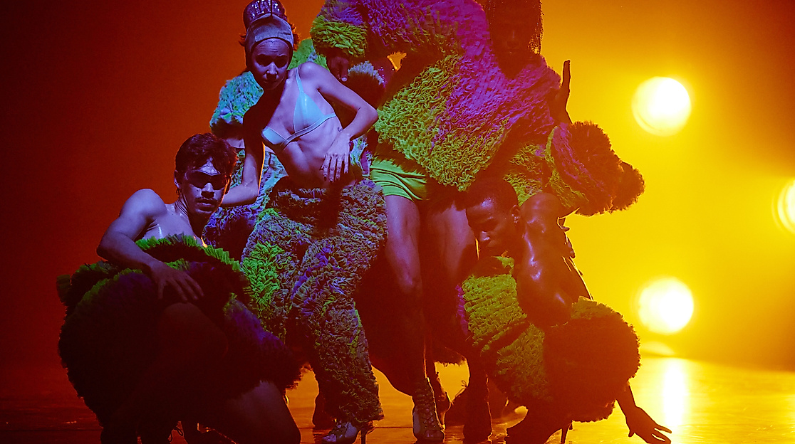 Ausschnitt der Produktion Trapicana. Mehrere Performer*innen stehen in einer tänzerischen Pose nah beieinander und werden von orangenem Licht angestrahlt.