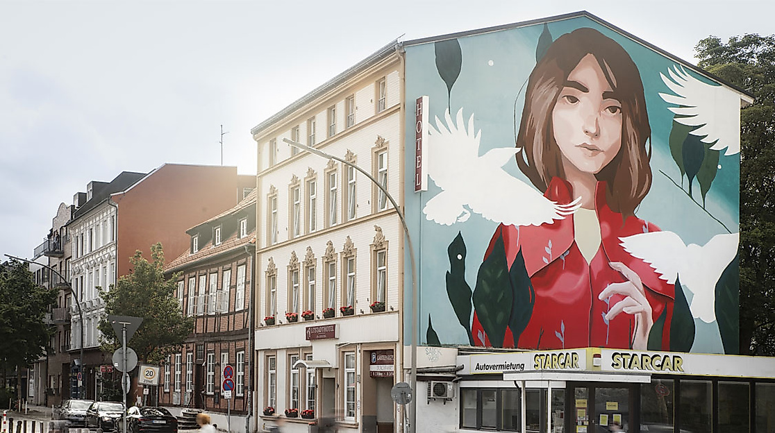 Hausfassade in Hamburg-Harburg mit großem Wandgemälde einer Frau in Rot und fliegenden weißen Tauben.