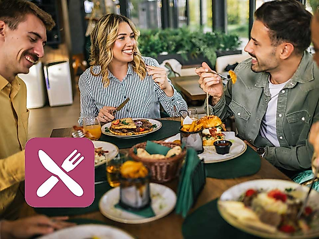Drei Personen sitzen an einem gedeckten Frühstückstisch in einem hellen, modern eingerichteten Café mit Blick ins Grüne. Die Stimmung ist lebhaft und freundlich – alle lächeln und unterhalten sich. Auf dem Tisch stehen liebevoll angerichtete Frühstücksteller mit Brötchen, Käse, Obst, Kuchen und Getränken wie Kaffee und Saft. Im Vordergrund ist ein Dessert mit Erdbeeren zu sehen. Links unten im Bild ist ein Icon mit Messer-und-Gabel-Symbol auf rosa Hintergrund eingeblendet, das auf ein kulinarisches Angebot hinweist. Die Szene vermittelt gemeinsames Genießen und den entspannten Start in den Tag – passend zum Schlemmer-Angebot der Hamburg CARD.