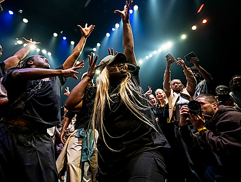 Eine dynamische Szene, bei der einen menge Menschen um eine Tänzerin herumstehen und sie mit Gesten anfeuern und jubeln. Die Tänzerin hebt streckt ihre Arme nach oben, spreizt die Finger. Rechts unten fotografiert jemand die Tänzerin von Nahem.