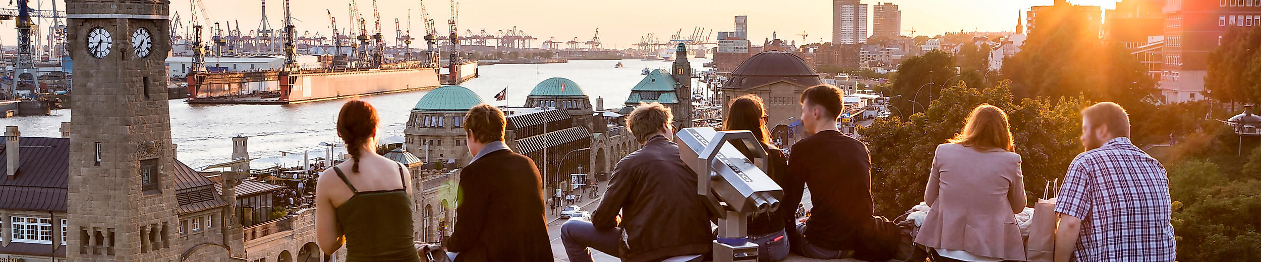 Menschen sitzen bei Sonnenuntergang oberhalb der Landungsbrücken mit Blick auf Hafen und Elbe