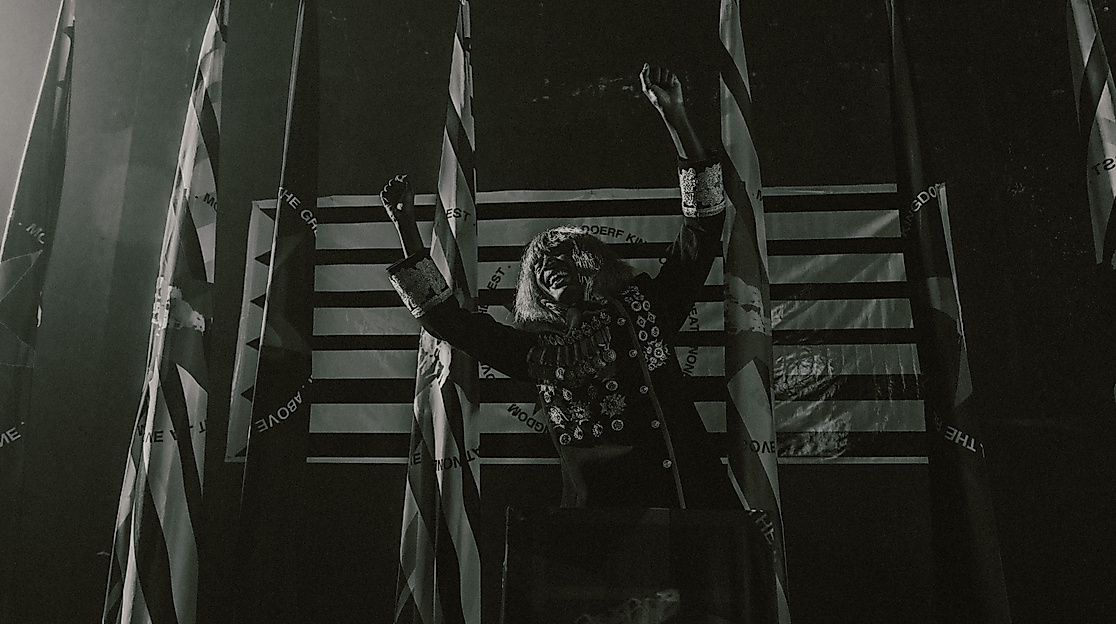 KABEAUSHÉ steht in einer Uniform mit vielen Orden auf einer Bühne vor vier Flaggen, streckt die Arme seitlich nach oben und lacht.