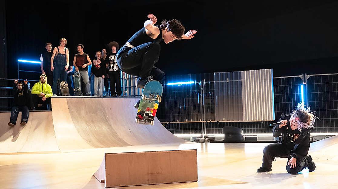 Mette Ingvartsen Skatepark Bea Borgers 21Eine Skateboarderin springt dynamisch über ein Hindernis, während eine Performerin mit Mikrofon in der Hocke singt und eine Gruppe Zuschauer:innen im Hintergrund zuschaut.