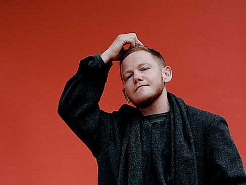 Kae Tempest steht in schwarz gekleidet vor einer bunten Wand und hat eine Hand auf den Kopf gelegt.