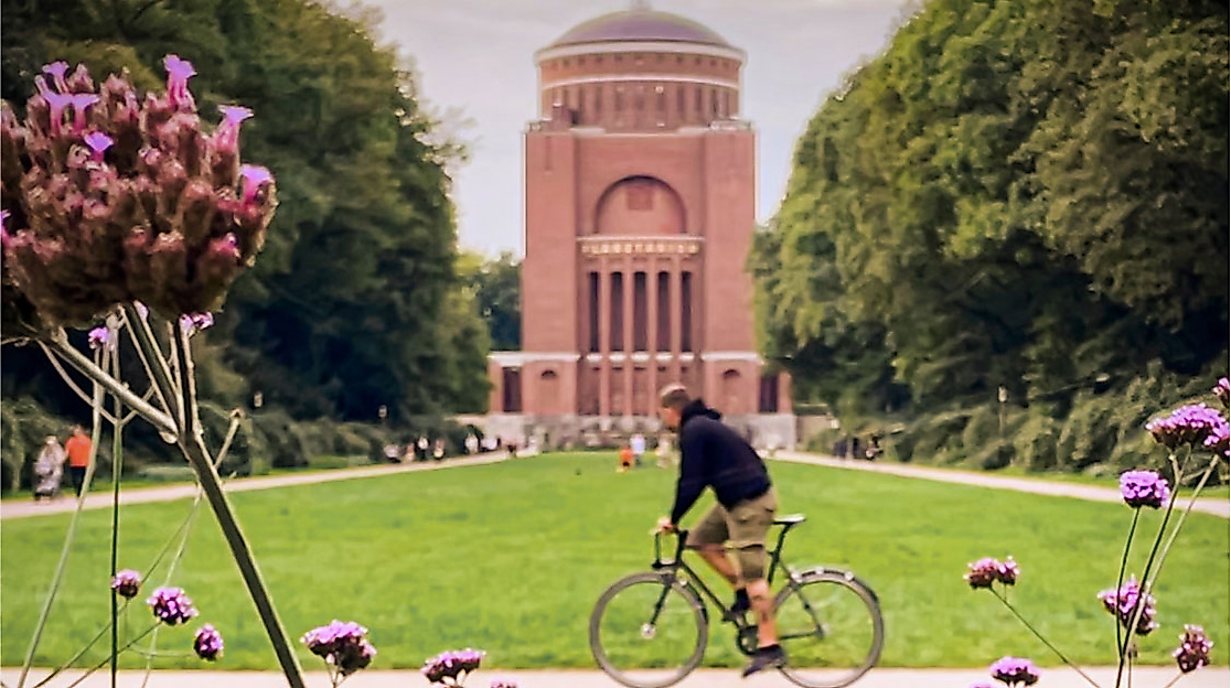 Radfahrer im Hamburger Stadtpark vor dem Planetarium, flankiert von Blumen und Baumallee