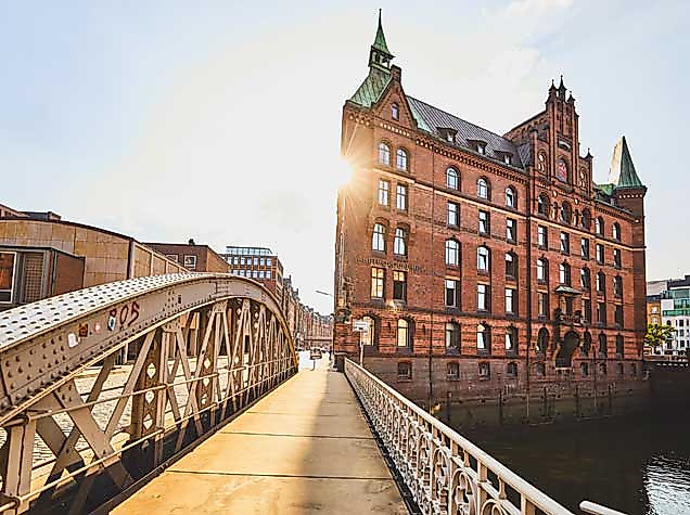 Ein sonniger Tag in der Hamburger Speicherstadt. Im Bildvordergrund führt eine historische Stahlbrücke mit Holzbohlen auf ein imposantes rotes Backsteingebäude mit Türmchen und Giebeln zu. Die Sonne scheint durch die Gebäudekante hindurch und erzeugt eine warme Lichtstimmung.