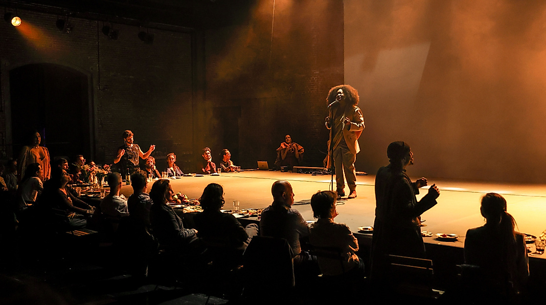 Ein stimmungsvolles Theaterbild: Menschen sitzen an einer langen Tafel voller Essen, während auf der Bühne eine Person mit Mikrofon spricht oder singt. Warmes Licht, intime Atmosphäre, Gemeinschaft.