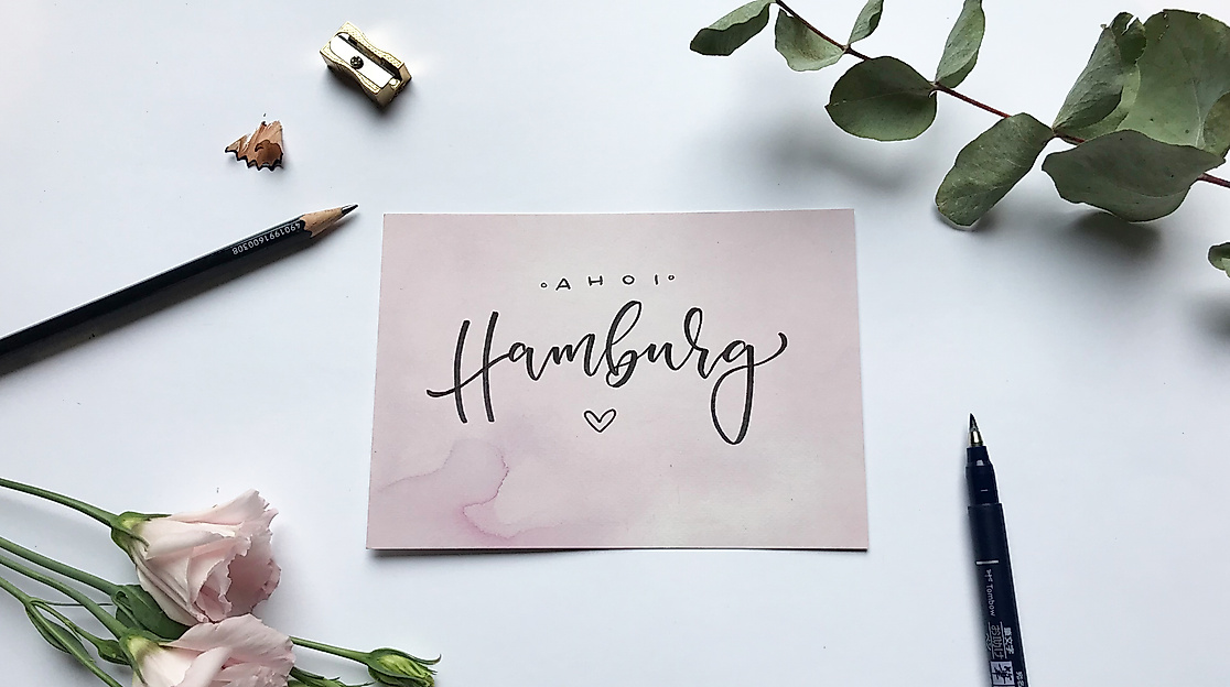 Karte mit Lettering-Schriftzug „Hamburg“ auf weißem Tisch, umgeben von Blumen, Stiften und Eukalyptus