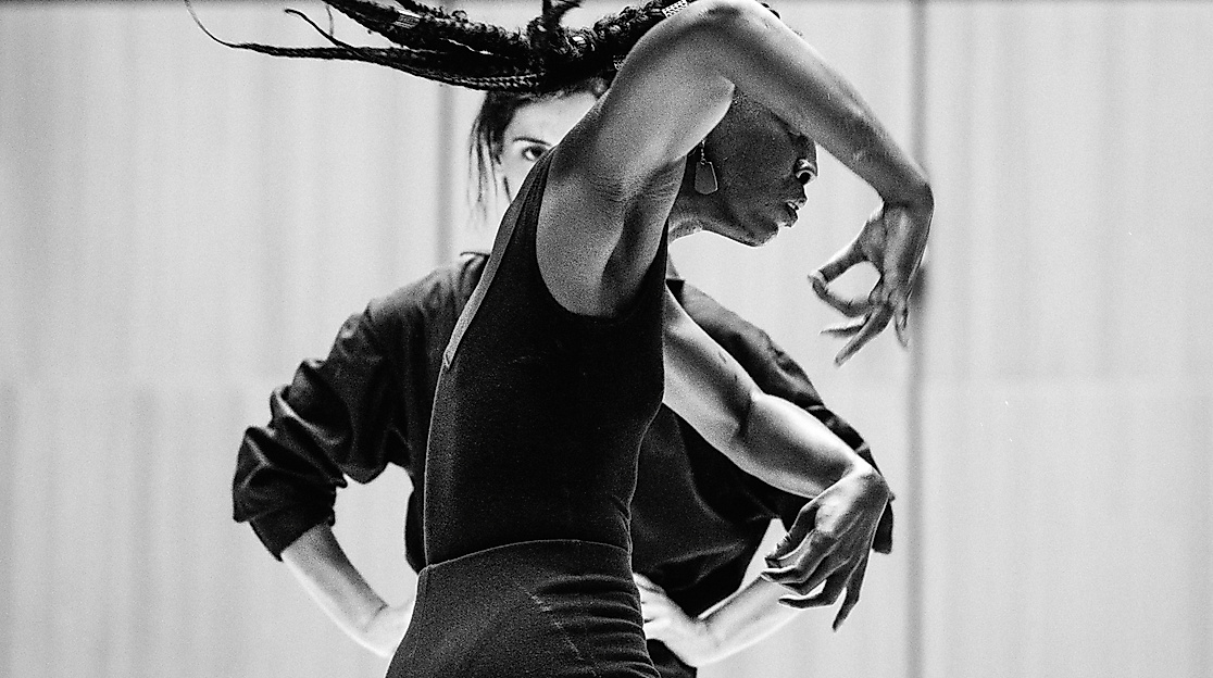 Eine Performerin ist von der Seite zu sehe, sie bewegt ihre Arme ausdrucksstark. Im Hintergund ist eine zweite Person zu sehen, die sie dabei beobachtet.