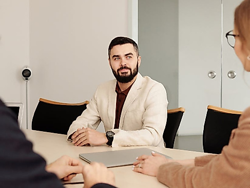 Ein Mann im weißen Jackett sitzt zwei weiteren Personen an einem Tisch gegenüber.