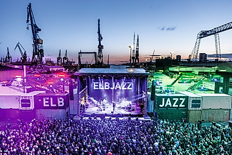 Beleuchtete Bühne des ELBJAZZ Festivals im Hamburger Hafen mit Menschenmenge und Hafenkränen bei Abenddämmerung.