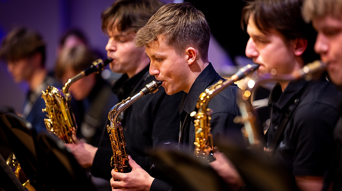 Ausschnitt eines Orchesters. Mehrere Personen stehen mit unterschiedlichen Blechblasinstrumenten nebeneinander und machen Musik.