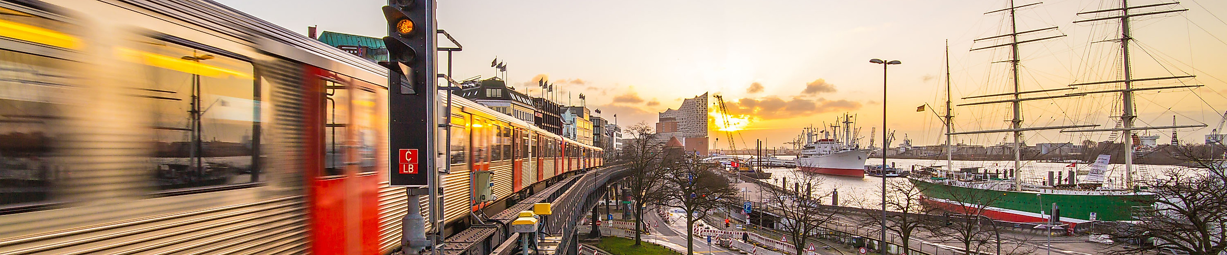 U-Bahn an den Landungsbrücken bei Sonnenuntergang, mit Blick auf Elbphilharmonie und Museumsschiffe.
