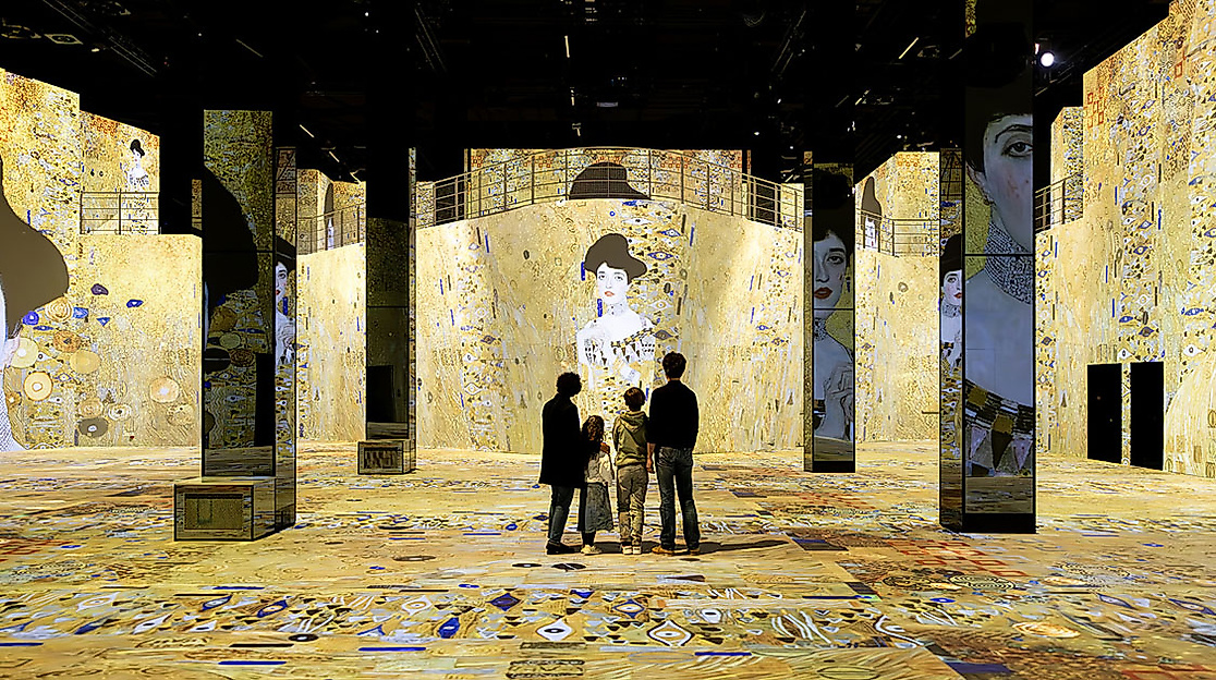 Besucher:innen in goldgelbem Raum mit Klimt-Projektionen in der immersiven Ausstellung im Port des Lumières Hamburg
