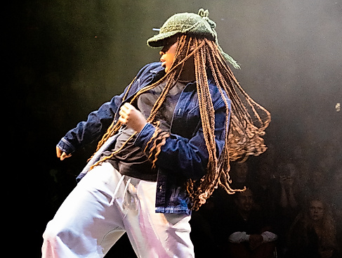 Eine Person mit Strickmütze, langen Braids und lockerer Kleidung steht auf einer Bühne und bewegt ihre Hüften mit gebeugten Knien in eine Richtung. Sie zieht sich mit dem gegenüberliegenden Arm in dieselbe Richtung hoch.