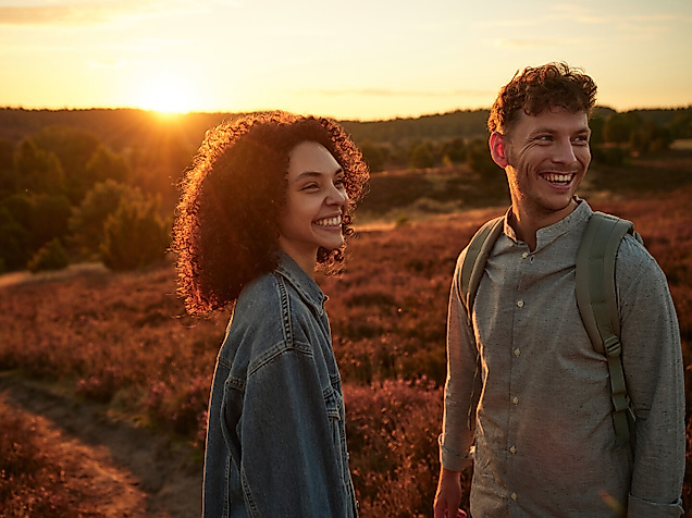 Ein junger Mann und eine junge Frau stehen im Sonnenuntergang lachend in der Lüneburger Heide, die lila blüht.