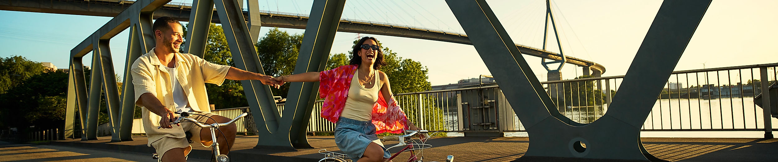 Zwei junge Leute fahren lachend mit dem Fahrrad über eine Brücke und halten sich dabei an den Händen.