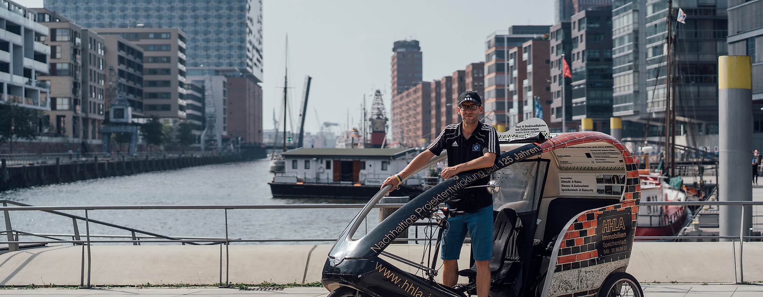 Rikscha-Fahrer mit Fahrgästen in der HafenCity, im Hintergrund die Elbphilharmonie und moderne Architektur.
