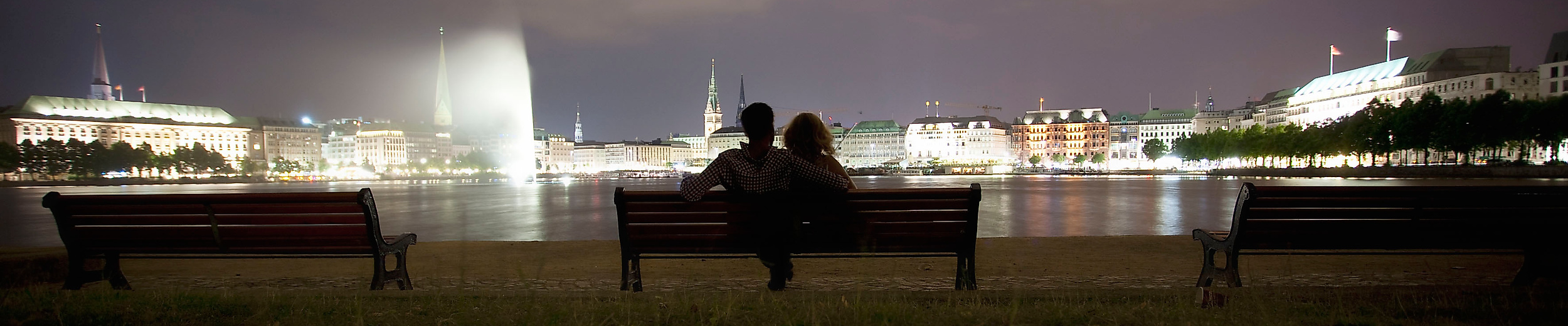 Nächtlicher Blick auf die Binnenalster mit Alsterfontäne und beleuchteter Skyline, Menschen sitzen auf einer Bank