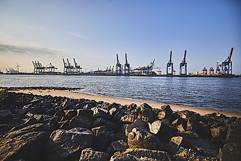 Blick vom Elbstrand auf Containerkräne im Hamburger Hafen bei Abendlicht, mit Steinen im Vordergrund