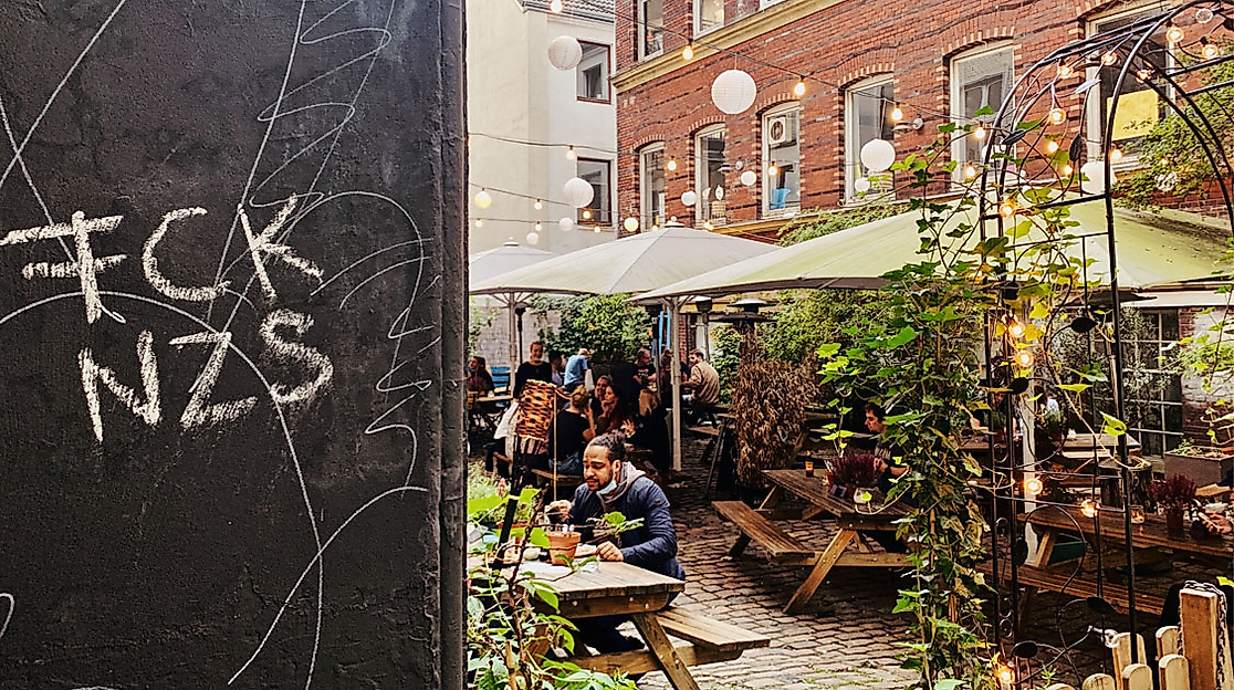 Belebter Innenhof mit Cafégarten im Hamburger Karolinenviertel; links politische Botschaft auf schwarzer Wand.