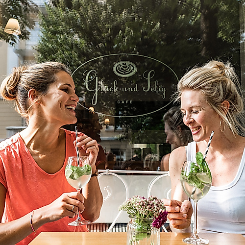 Zwei Personen stoßen im Café in entspannter, sommerlicher Atmosphäre an.