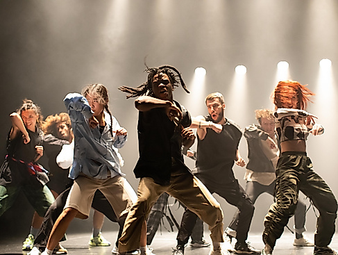 Eine energiegeladene Tanzgruppe führt auf einer Bühne unter dramatischer Beleuchtung eine kraftvolle Choreografie auf.