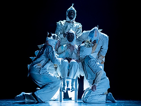 Vier Performer*innen stehen dicht neben einer auf einem Stuhl sitzenden Person. Alle tragen helle Kostüme mit großen Zacken auf dem Rücken.