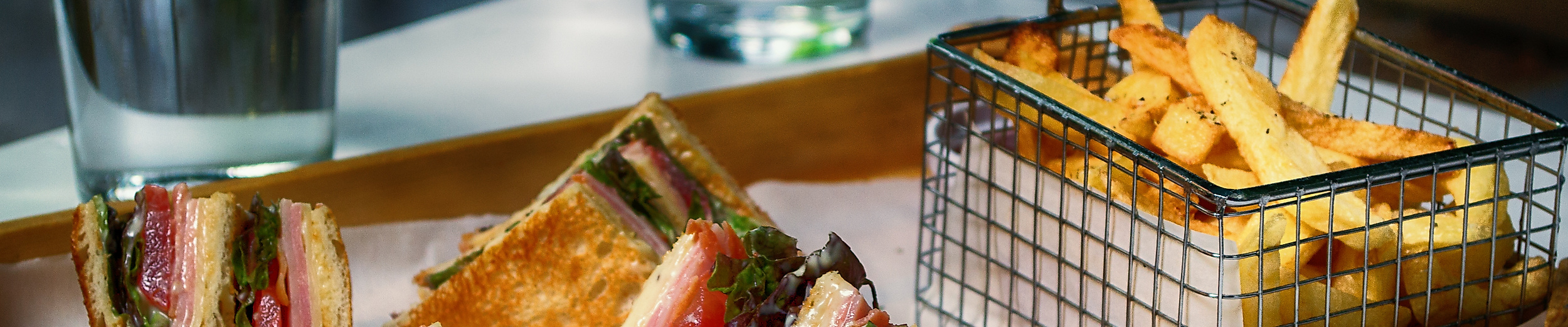 Toast-Sandwiches mit Pommes in Metallkörbchen auf Holztablett, dazu zwei Gläser Wasser.