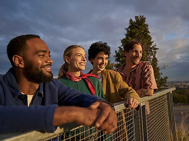 Vier junge Erwachsene stehen entspannt an einem Geländer und blicken lächelnd in die Ferne. Die Szene spielt im warmen Licht eines Sonnenuntergangs, im Hintergrund sind Bäume und ein weiter Blick über die Stadt Hamburg zu erkennen. Die Gruppe wirkt gelöst und fröhlich – ein Moment gemeinsamer Entdeckung und Lebensfreude