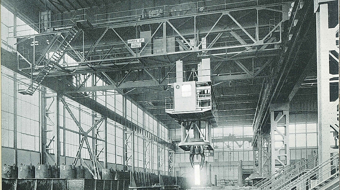Ein altes schwarz-weiß Foto der ehemaligen Fabrikhallen von Kampnagel mit Gerüsten, einem Kran und aufgereihten Metall-Tonnen.