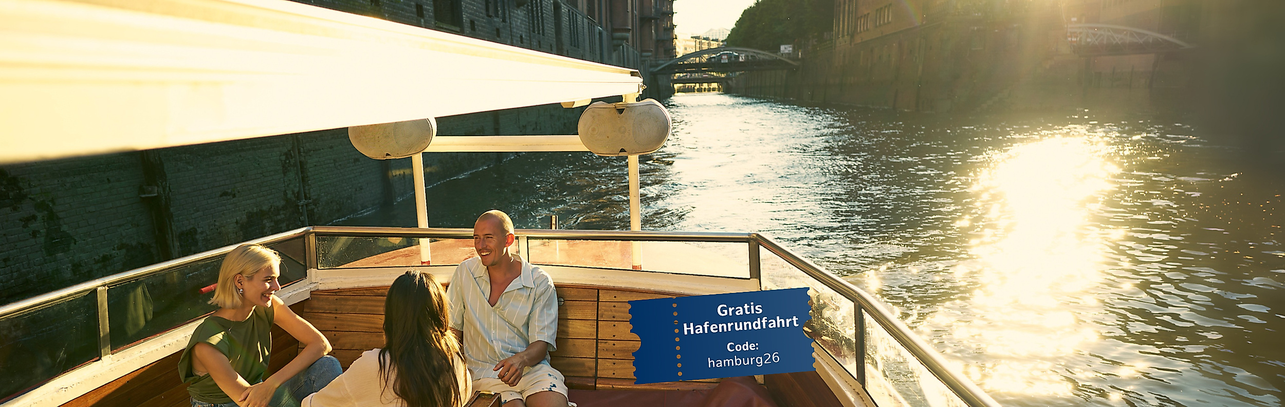 Drei Personen auf einer Barkasse bei Sonnenuntergang, Speicherstadt im Hintergrund, Hinweis auf "Gratis Hafenrundfahrt mit Code: hamburg2026"