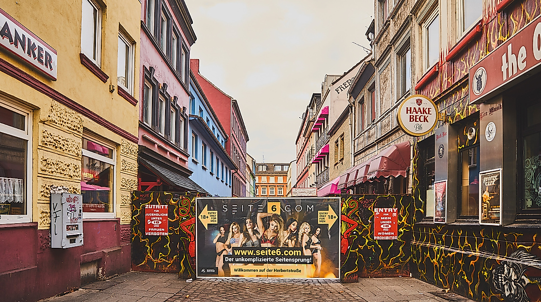 Blick in die Herbertstraße mit Sichtschutzwand, flankiert von bunt beleuchteten Fassaden und Schildern auf der Reeperbahn