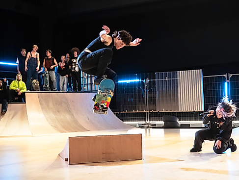Mette Ingvartsen Skatepark Bea Borgers 21Eine Skateboarderin springt dynamisch über ein Hindernis, während eine Performerin mit Mikrofon in der Hocke singt und eine Gruppe Zuschauer:innen im Hintergrund zuschaut.