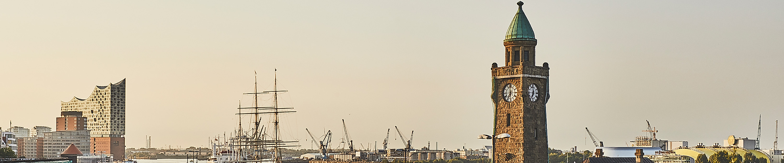 Landungsbrücken mit Uhrenturm im Morgenlicht, Hafenkräne und Elbe im Hintergrund beim Sonnenaufgang