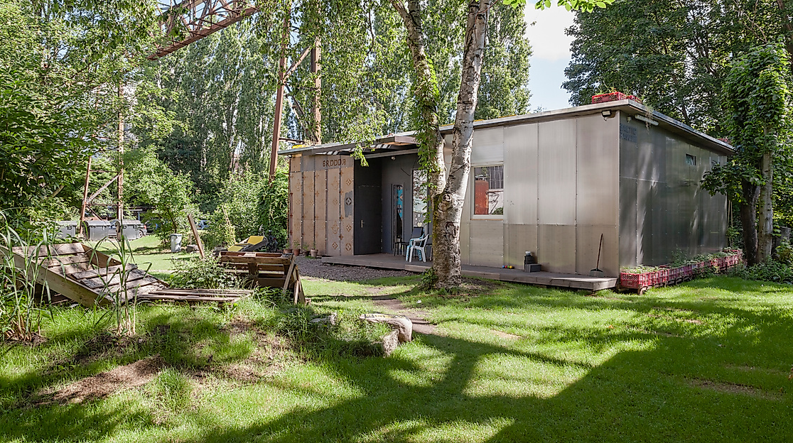Foto vom Migrantpolitan auf Kampnagel. Ein kleines einstöckiges Haus mit einem grünen Garten, Bänken aus Holzpaletten, umsäumt von vielen Bäumen.