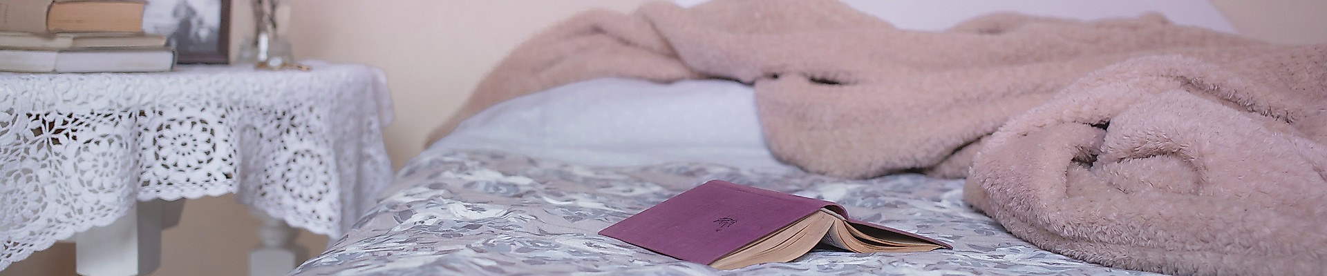 Offenes Buch liegt auf gemachtem Bett mit rosa Decke, daneben Nachttisch mit Büchern und Trockenblumen.