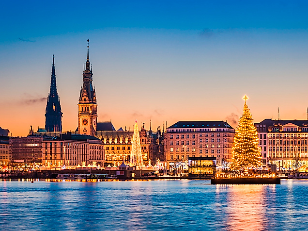 Weihnachtsbaum auf der Alster vor dem beleuchteten Jungfernstieg und der Hamburger Altstadtsilhouette bei Sonnenuntergang.