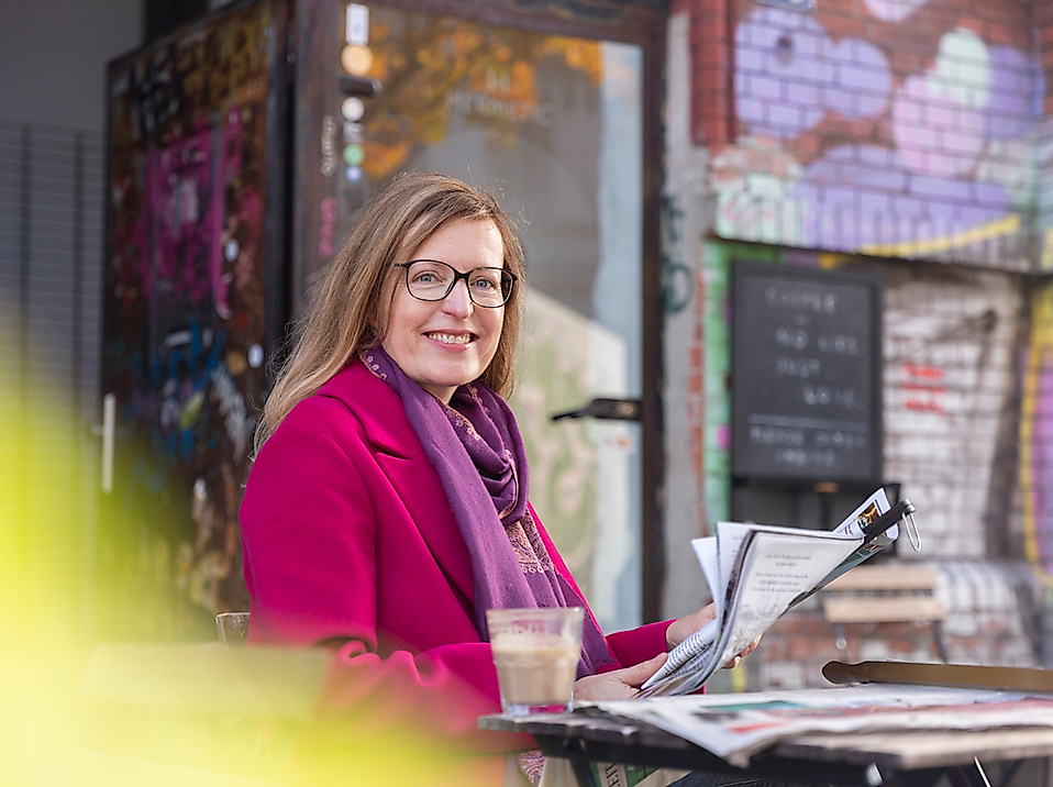 Lächelnde Frau mit Zeitschrift und Getränk im Straßencafé vor bunt besprühter Wand im Hamburger Schanzenviertel.