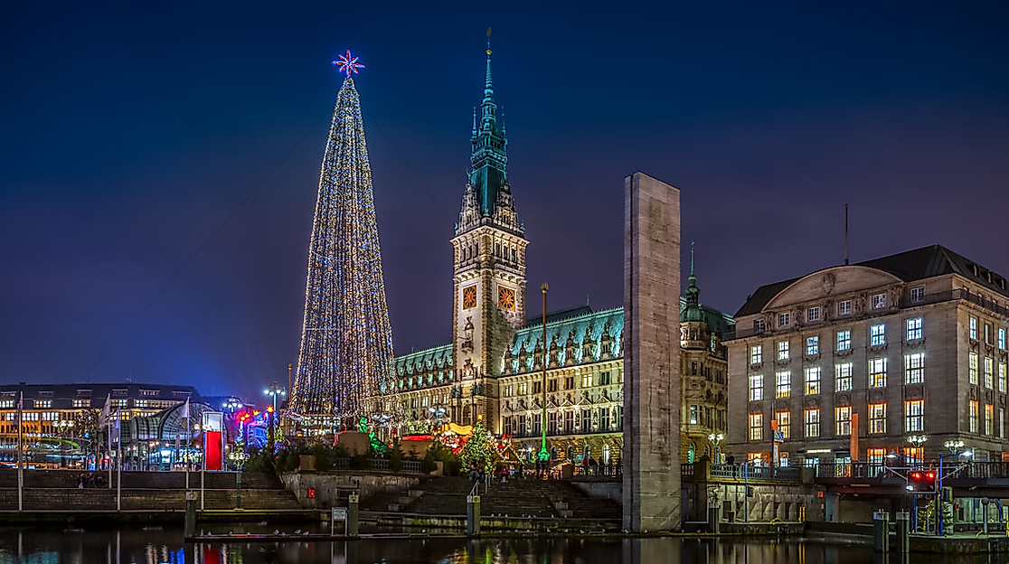 Festlich beleuchtetes Hamburger Rathaus und Weihnachtsmarkt bei Nacht spiegeln sich im Alsterfleet.