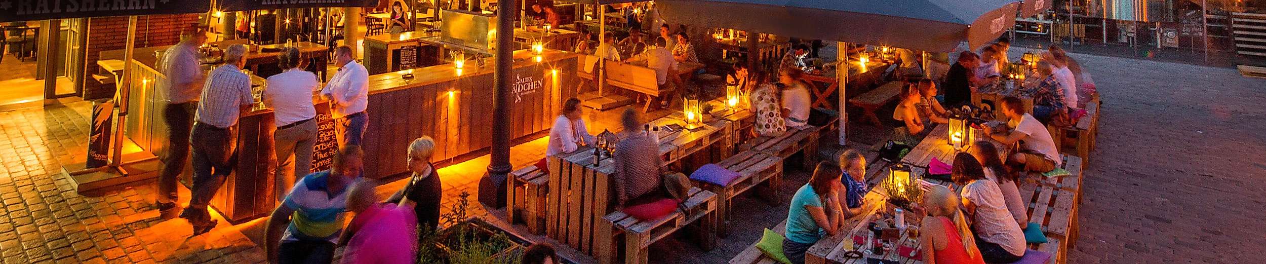 Belebter Außenbereich mit Bars und Gästen am Abend, im Hintergrund der Hamburger Fernsehturm