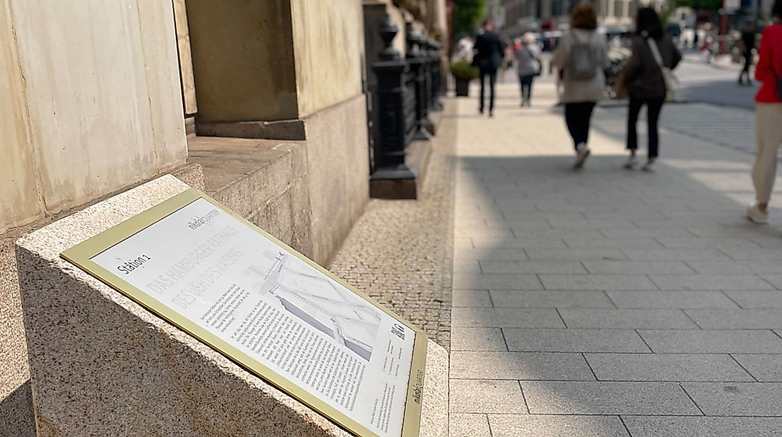 Tastbare Infotafel Nikolaiquartier in Hamburg an einem historischen Gebäude, vorbeigehende Passant:innen auf breitem Gehweg