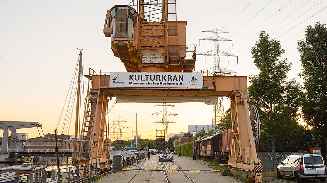 Gelber Kulturkran in Hamburg-Harburg bei Sonnenuntergang, umgeben von Autos, Bahngleisen und Hafenatmosphäre