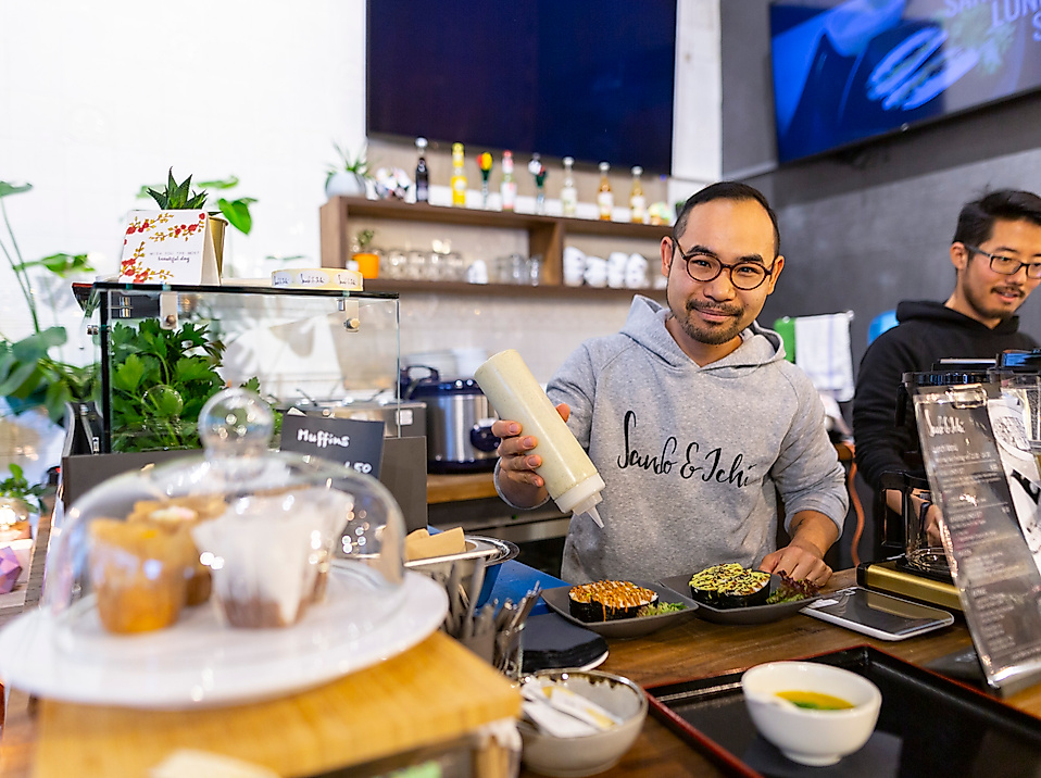 Zwei Köche bereiten asiatische Speisen an einer modernen Theke zu, im Vordergrund Kuchen unter Glasglocke
