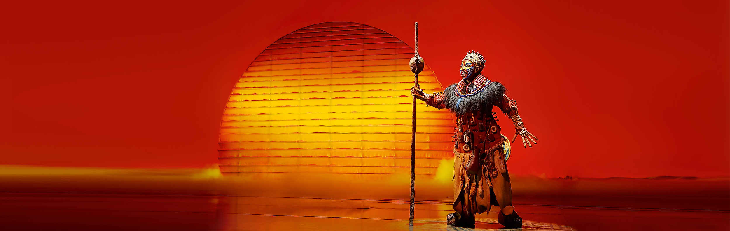 Darstellerin in aufwendigem Kostüm vor roter Kulisse im Musical Der König der Löwen in Hamburg.